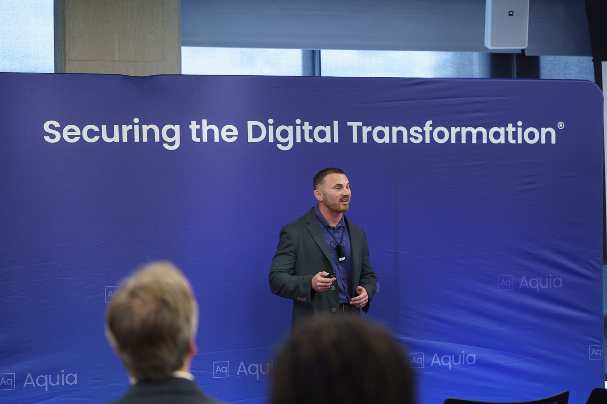 Chris Hughes standing in front of a blue banner that says Securing the Digital Transformation. He is wearing a microphone and holding a slide clicker as he delivers the opening remarks for the event.