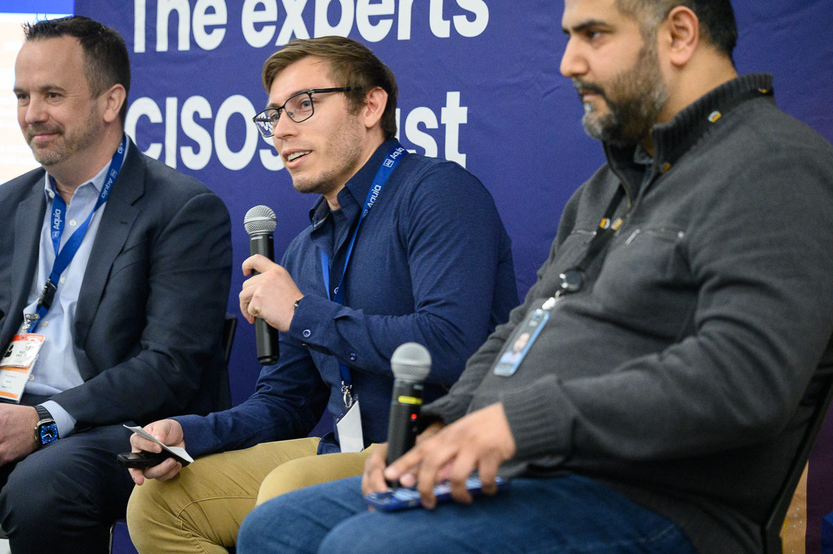 Close-up of three men in front of a blue banner that says The Experts CISOs Trust. Lloyd Evans is in the center speaking into a microphone.